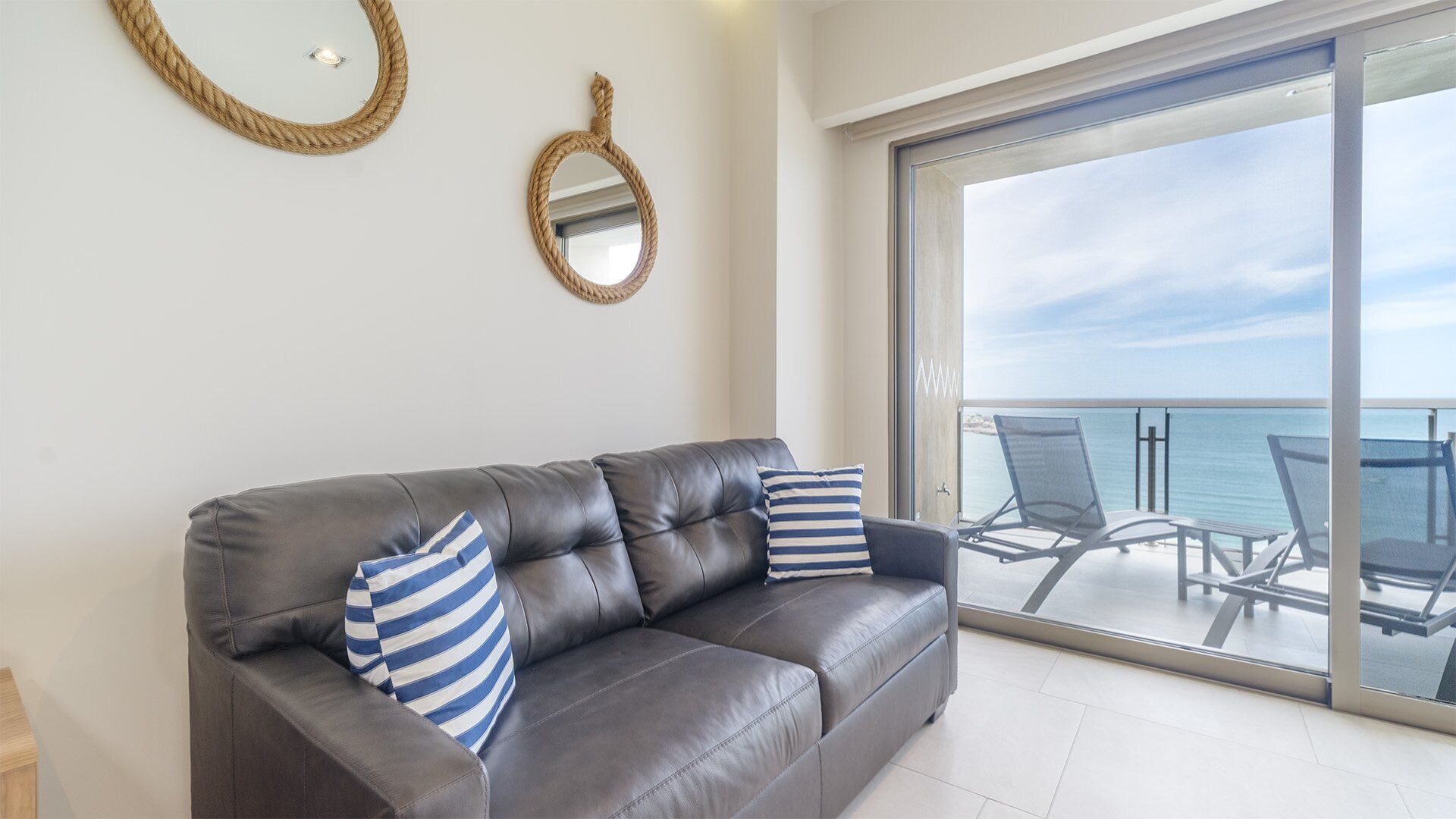 Un canapé en cuir foncé agrémenté de coussins à rayures, un miroir rond et un balcon avec des chaises d’où l’on peut admirer la mer.