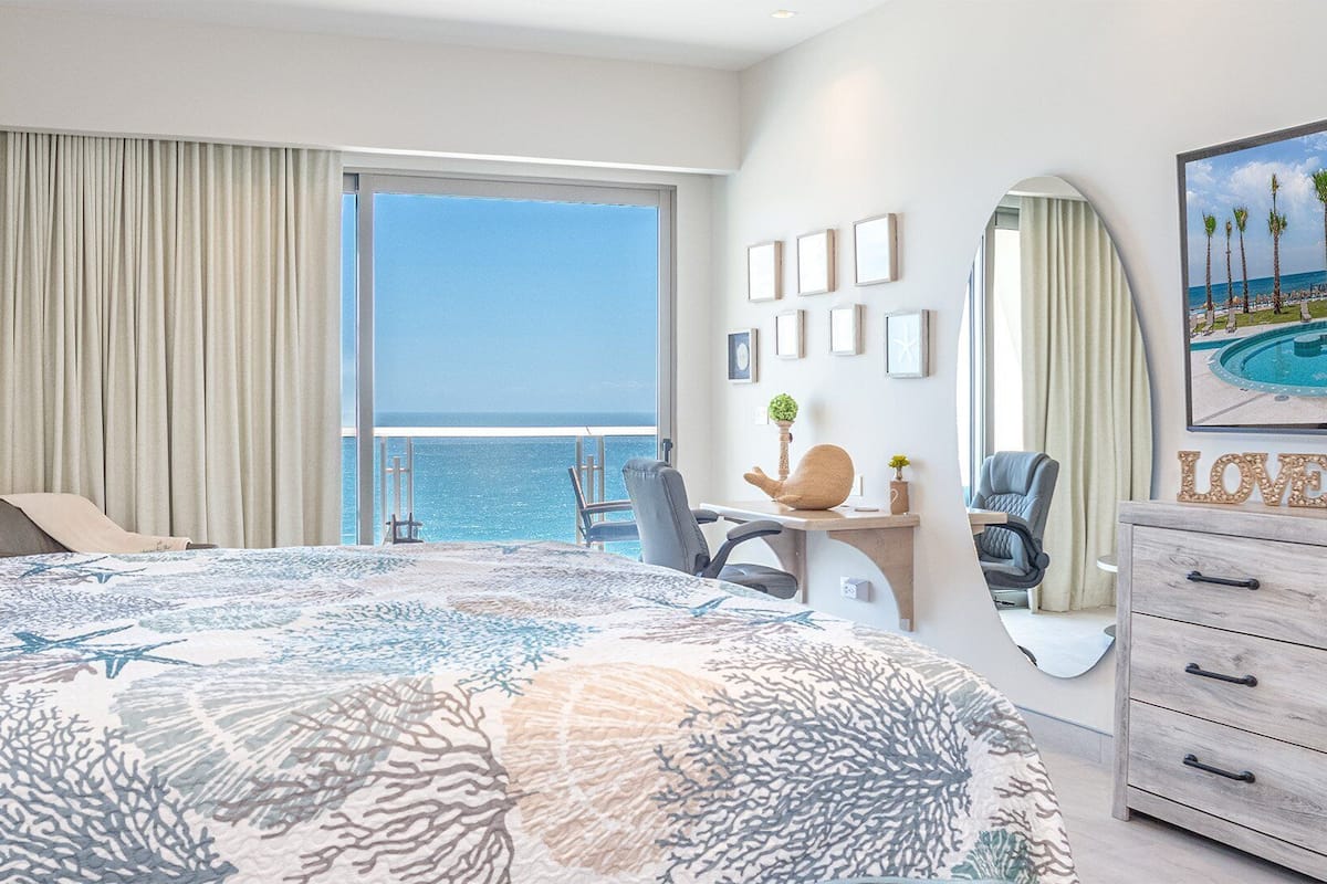 Ein Hotelzimmer mit einem großen Bett, einem Schreibtisch, einem Sessel, einer Kommode und einem großen Fenster mit Blick aufs Meer.