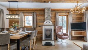 A cozy living room with a stone fireplace, wooden beams, a dining area with a wooden table and chairs, and a television on a wooden stand. - Chalet on the Katschberg with sauna (Katschberg)