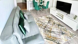 A modern living room with a grey sofa, a dining area with green chairs, a TV on a white stand, and a large abstract rug. - Penthouse Apartment in Portsmouth: Close to Beach (Portsmouth)