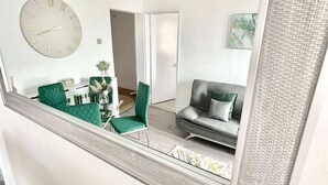A modern living room with a large wall clock, a sofa, and a dining area with green chairs. - Penthouse Apartment in Portsmouth: Close to Beach (Portsmouth)