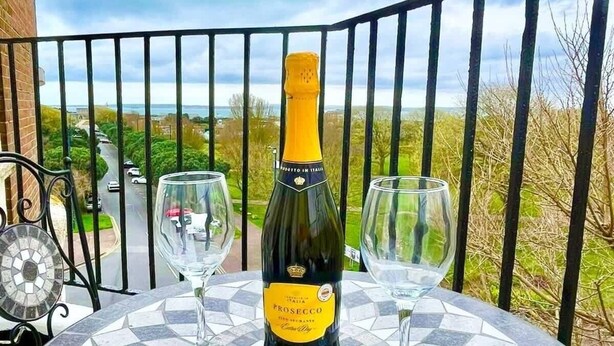 A round table with a mosaic tile design, a bottle of Prosecco, and two wine glasses on a balcony with a metal railing and a view of greenery and a road. - Penthouse Apartment in Portsmouth: Close to Beach (Portsmouth)