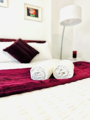 A neatly made bed with a red blanket and two rolled towels, a white bedside lamp, and a dark red pillow.