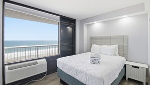 A bedroom with a large window overlooking a beach, a bed with white bedding, and a white bedside table. - Beachfront Studio | Kitchen + Pool & Picnic Area (Ormond Beach)