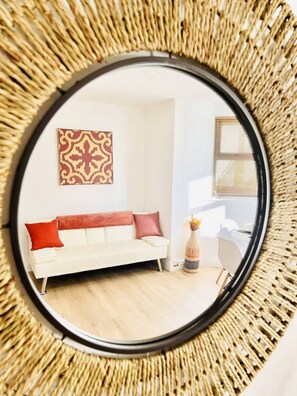 Un miroir rond doté d’un cadre tressé, qui reflète une pièce comprenant un canapé blanc, des coussins rouges, une décoration murale à motifs et une petite plante.