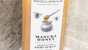 Un pot de gommage corporel au miel de Manuka de la marque Russell & Windsor, doté d’un distributeur à pompe, posé sur une surface blanche devant un mur texturé.