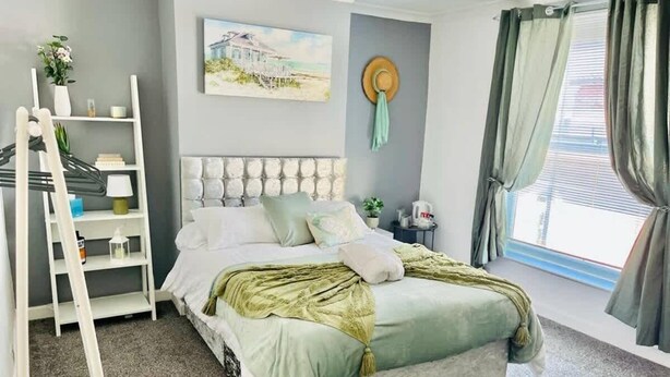 Une chambre à coucher avec un lit, une échelle blanche, une étagère avec des objets, un tableau au mur et une fenêtre avec des rideaux.