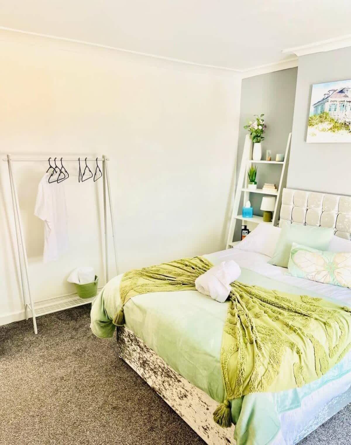 A bedroom with a bed, a green and white bedspread, a white towel, a green basket, and a white towel rack.