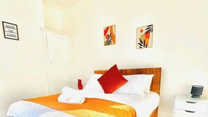Ein ordentlich bezogenes Bett mit einem roten Kissen, weißer Bettwäsche und einer dekorativen orangefarbenen Decke. Ein weißer Nachttisch mit einer Lampe und einer kleinen Vase steht daneben.