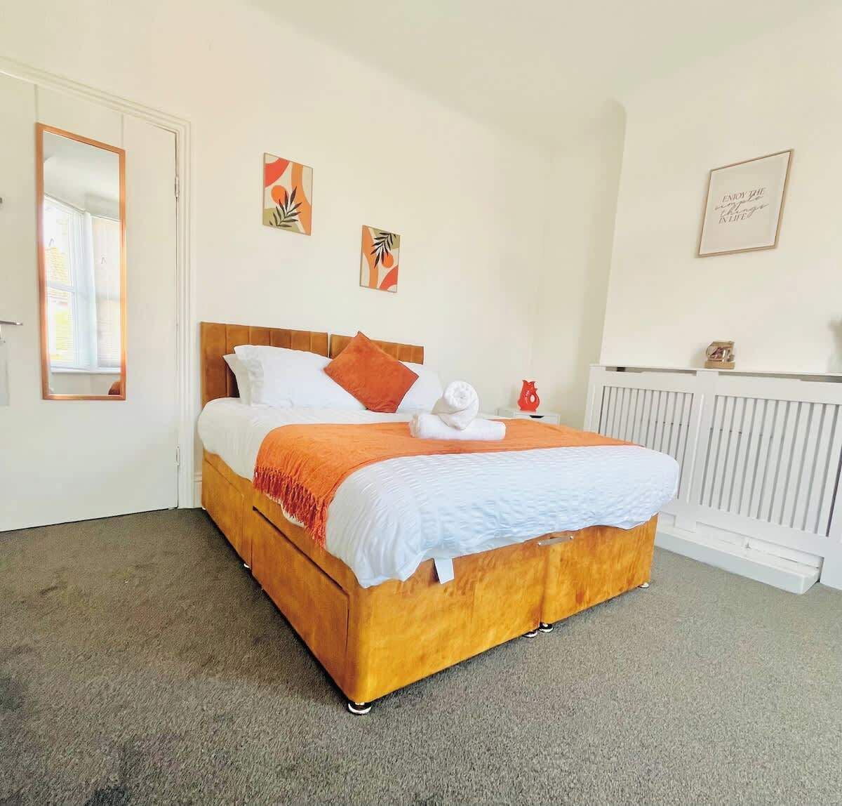 Un lit bien fait, avec une tête de lit en bois, une literie blanche et un plaid orange. Deux œuvres d’art sont accrochées au mur, ainsi qu’une citation encadrée.