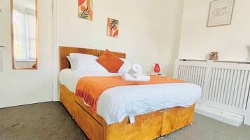 Un lit bien fait, avec une tête de lit en bois, une literie blanche et un plaid orange. Deux œuvres d’art sont accrochées au mur, ainsi qu’une citation encadrée.