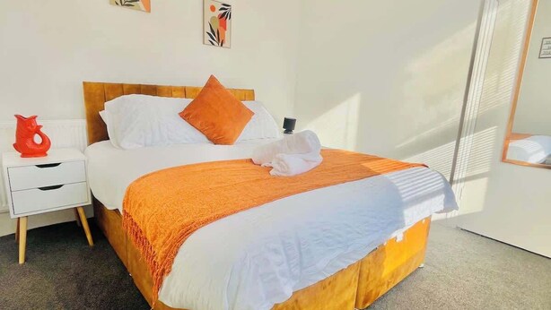 Ein Hotelzimmer mit einem hölzernen Bett, weißer Bettwäsche, einer orangefarbenen Decke und einem weißen Nachttisch mit einer roten Vase.