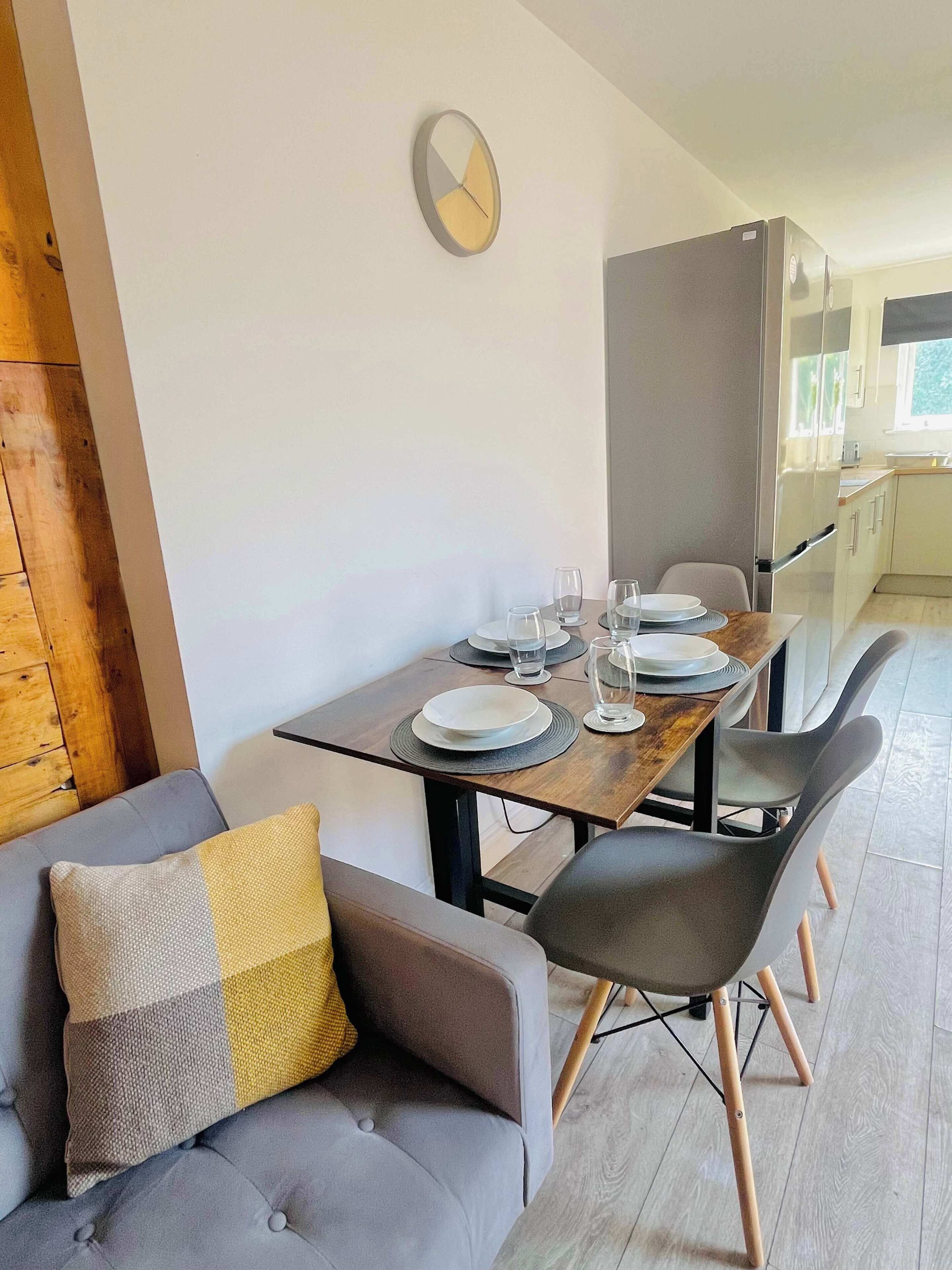 Un'area pranzo moderna con un tavolo di legno apparecchiato per quattro persone, un divano grigio con un cuscino giallo e una cucina sullo sfondo.