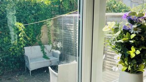 A window with a view of a garden, a white chair, and a potted plant with flowers.