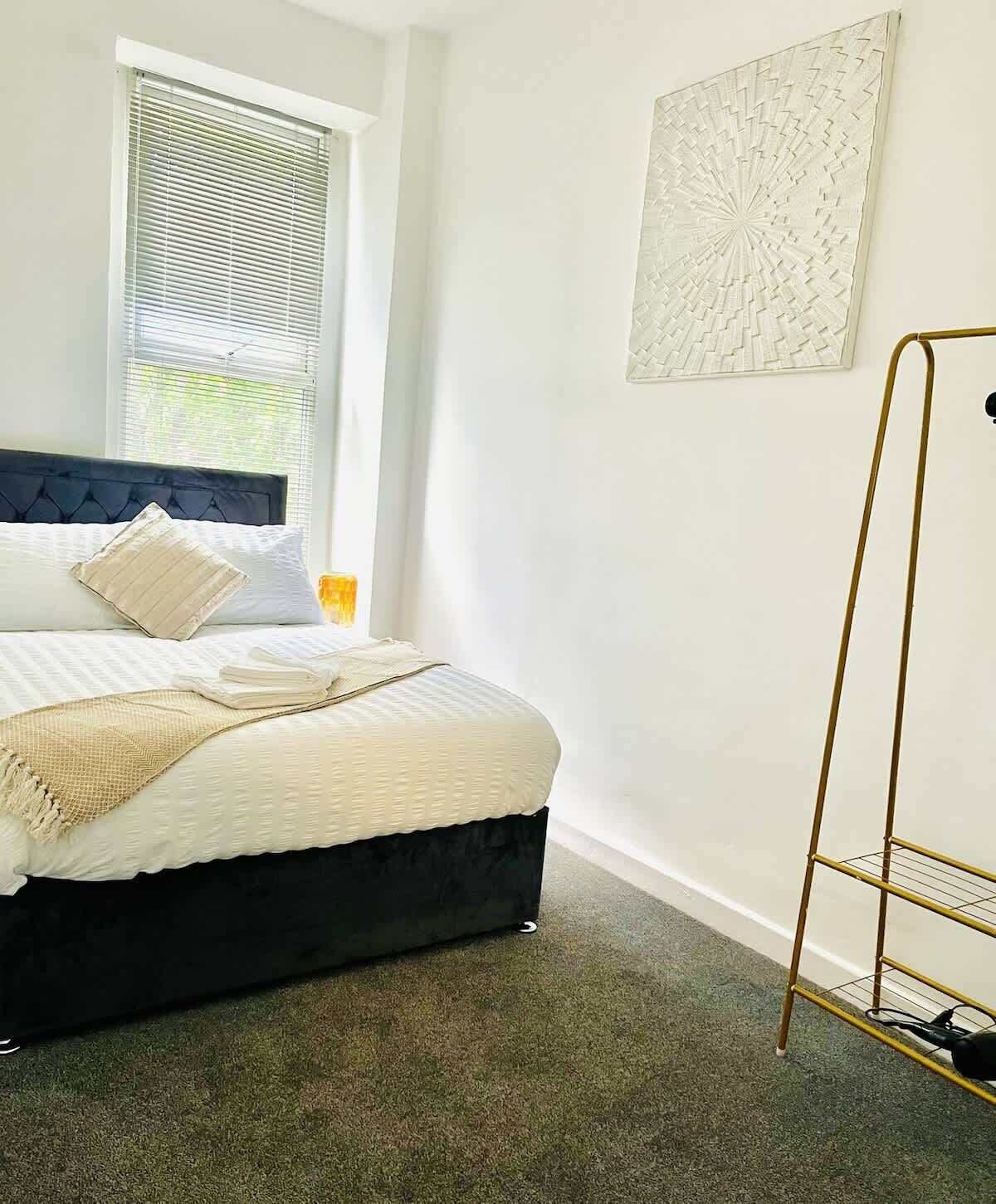 A bedroom with a bed, a window with blinds, a bedside table with a lamp, and a wall-mounted artwork.