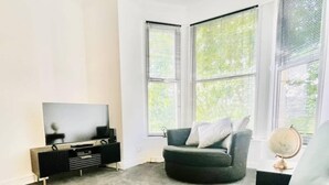 A modern living room with a grey sofa, a black entertainment unit, and a large window with blinds. - Portsmouth Apartment - Seaside Gem (Portsmouth)