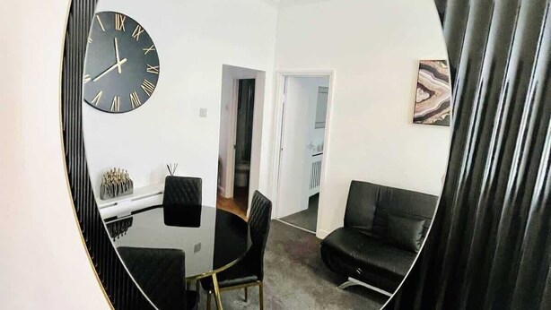 A room with a round mirror reflecting a dining area, a black sofa, and a clock on the wall. - Portsmouth Apartment - Seaside Gem (Portsmouth)