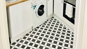 Een kleine keuken met witte kastjes, een wasmachine, een broodrooster en een klok aan de muur.