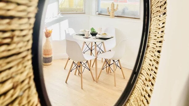 Een ronde spiegel met een gevlochten lijst toont een eethoek met een witte tafel, stoelen en een cactusdecoratie.
