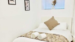 Een opgemaakt bed met witte lakens en een bruin kussen, een klein raam met jaloezieën en twee ingelijste foto's aan de muur.