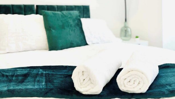 A neatly made bed with white linens and a teal throw blanket, a green pillow, and a white pillow. A lamp is on the nightstand beside the bed. - Clarence Road: Cozy King Room in Portsmouth (Portsmouth)