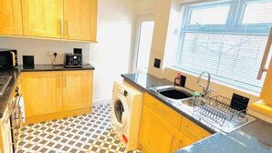 A compact kitchen with wooden cabinets, a washing machine, a sink with a dish rack, and a patterned floor. - Clarence Road: Cozy King Room in Portsmouth (Portsmouth)