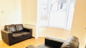 A living room with a black leather sofa, a smaller black ottoman, and a window with blinds. - Clarence Road: Cozy King Room in Portsmouth (Portsmouth)