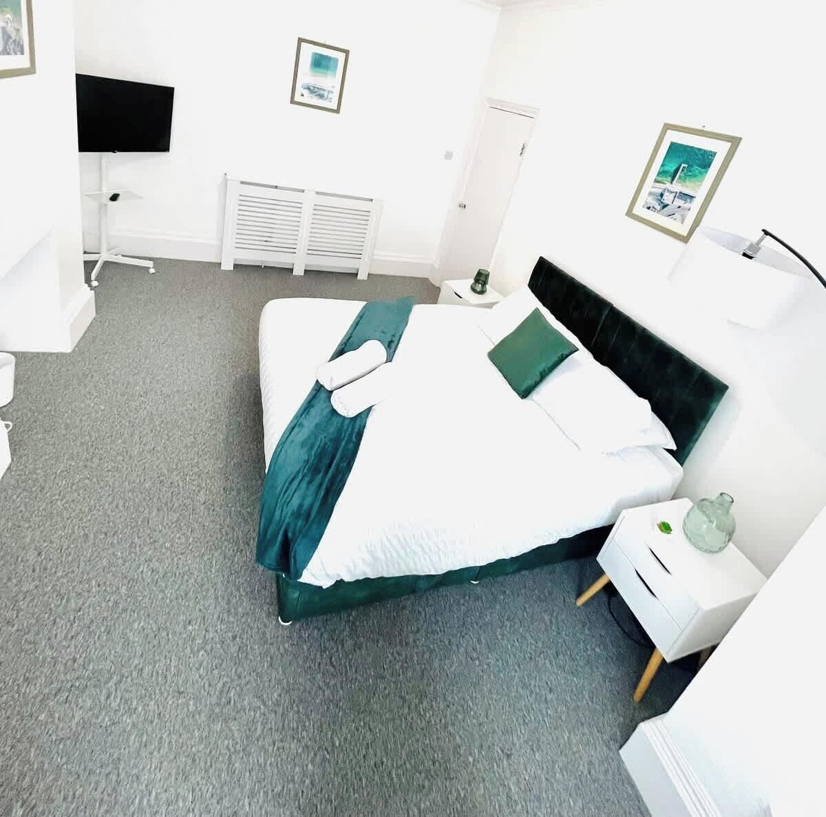 A neatly arranged hotel room with a bed, a bedside table, a chair, a radiator, a television, and framed pictures on the wall.