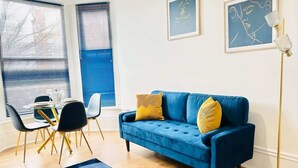 A modern living room with a blue sofa, yellow cushions, a black rug, and a dining area with a table and chairs. - Portsmouth Apartment: Cozy & Convenient Location (Portsmouth)