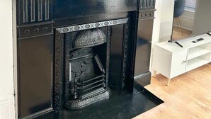 A black fireplace with ornate detailing, a white console table, and a mirror with a wavy gold frame. - Portsmouth Apartment: Cozy & Convenient Location (Portsmouth)