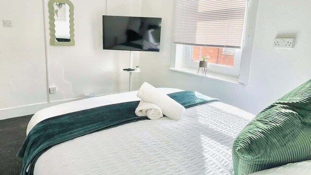 Une chambre d’hôtel avec un lit, un téléviseur à écran plat fixé au mur, une fenêtre avec des stores et un luminaire suspendu vert.