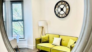 Ein Zimmer mit einem grünen Sofa, einer Wanduhr, einer Stehlampe und einem Fenster mit Jalousien.