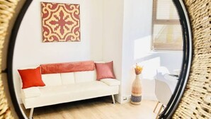 Ein runder Spiegel mit gewebtem Rahmen, der einen Raum mit einem weißen Sofa, roten Kissen, einem gemusterten Wandbehang und einer kleinen Pflanze zeigt.