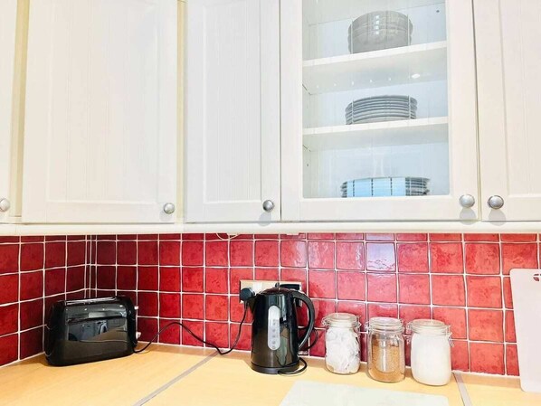 Une cuisine avec des meubles blancs, un plan de travail recouvert de carreaux rouges, un grille-pain noir, une machine à café et des bocaux de sucre et de farine posés sur le comptoir.