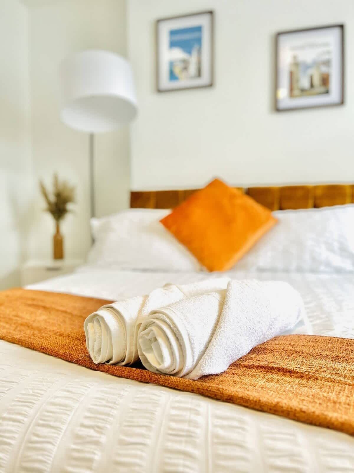 A neatly made bed with a folded white towel, a brown throw blanket, and an orange pillow.