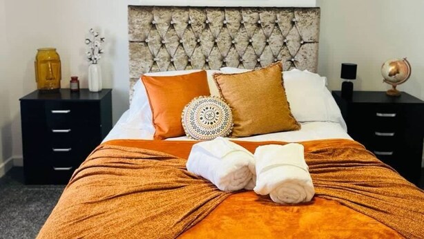 A neatly made bed with an orange blanket, two pillows, and two rolled towels. There is a headboard with a tufted design, and a small round mirror above the bed. - Cozy Room in Fratton: Featuring Hot Tub, Kitchen & Free WiFi (Fratton)