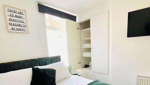 A neatly made bed with white and green bedding, a bedside table with a lamp, and a wall-mounted shelf with folded clothes. - Cleveland Road House: Garden, Kitchen & Free Parking (Portsmouth)