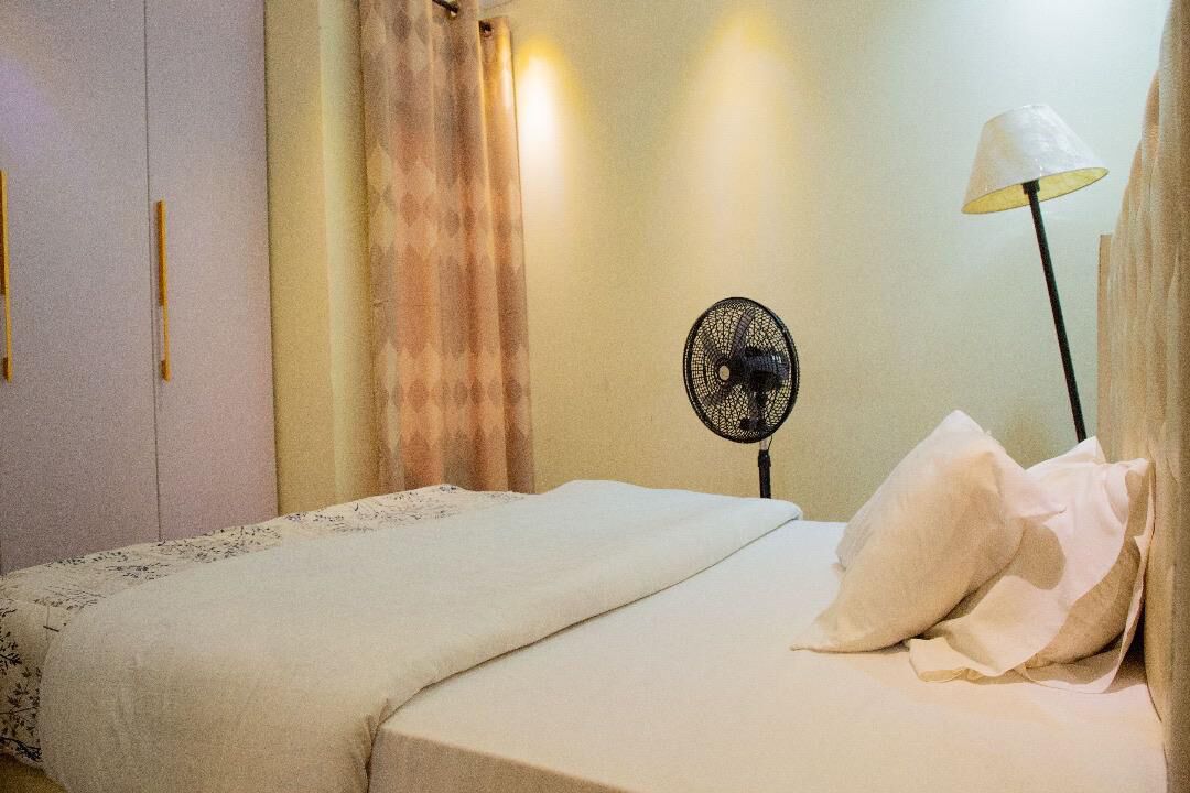 A bedroom with a bed, a standing fan, a lamp, and a curtain.
