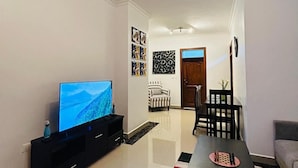 A modern living room with a flat-screen TV on a black stand, a patterned rug, a dining table with chairs, and a chandelier. - VYREX RESIDENCE (Kigali)