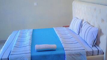 A neatly made bed with a blue blanket and striped pillows, a white headboard, and red slippers on the floor.