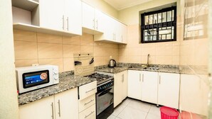 A compact kitchen with white cabinets, a microwave, a gas stove, and a window with bars. - VYREX RESIDENCE (Kigali)