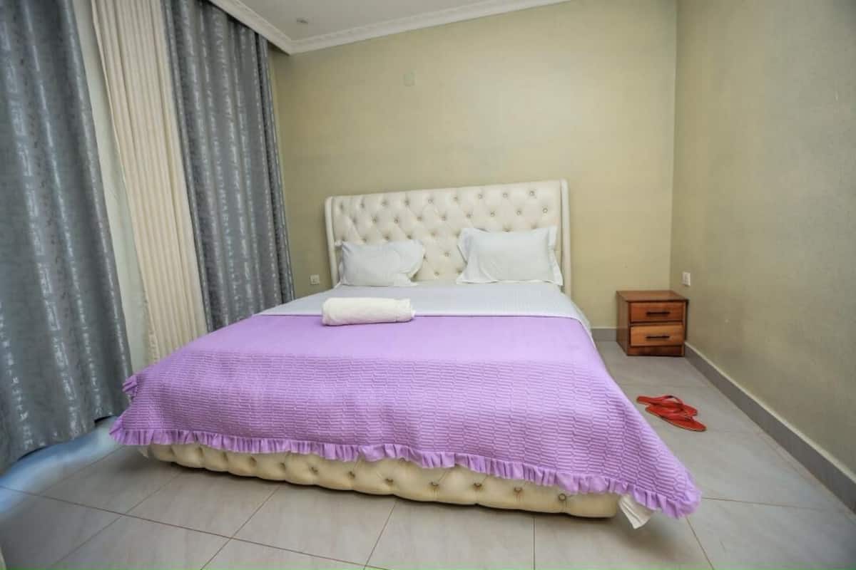 Una cama bien hecha con una colcha morada y una almohada blanca, una mesita de noche de madera con un cajón, y un par de pantuflas rojas sobre el suelo.