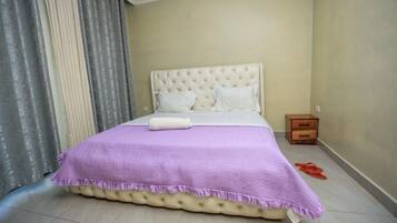 A neatly made bed with a purple bedspread and a white pillow, a wooden nightstand with a drawer, and a pair of red slippers on the floor.