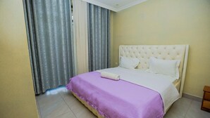 A hotel room with a bed, curtains, and a small wooden side table. - VYREX RESIDENCE (Kigali)