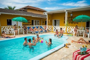 A swimming pool area with lounge chairs and umbrellas, surrounded by a yellow building with a white fence.