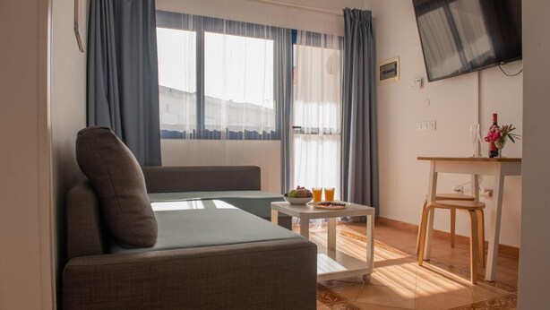 A modern living room with a sofa, a small table with fruit and drinks, a TV, and a window with curtains. - Casita Lola - Sunny Apartment in the closed complex with a swimming pool. (Costa Calma)