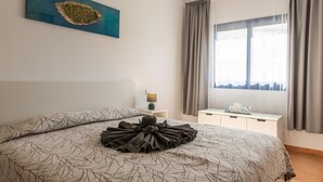 A bedroom with a bed, a nightstand, a lamp, a window with curtains, and a painting on the wall. - Casita Lola - Sunny Apartment in the closed complex with a swimming pool. (Costa Calma)