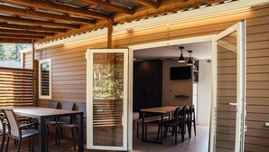 Un patio cubierto con piso de madera, una mesa de comedor con sillas y una puerta corrediza de vidrio que da a un espacio interior con televisor.