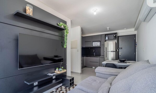 A modern living room with a flat-screen TV, a grey sofa, and a kitchen area with a fridge and microwave. - Comfort and Convenience Just 850m From Tambau Beach - Onws (João Pessoa)