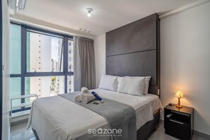 Ein modernes Hotelzimmer mit einem großen Bett, einem Nachttisch mit Lampe und Blick auf die Stadt durch ein großes Fenster.